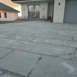 Ułożona szara kostka brukowa przed nowoczesnym domem jednorodzinnym z białymi ścianami i szarą bramą garażową; widoczne białe krzesła i stół na tarasie oraz dekoracje halloweenowe.