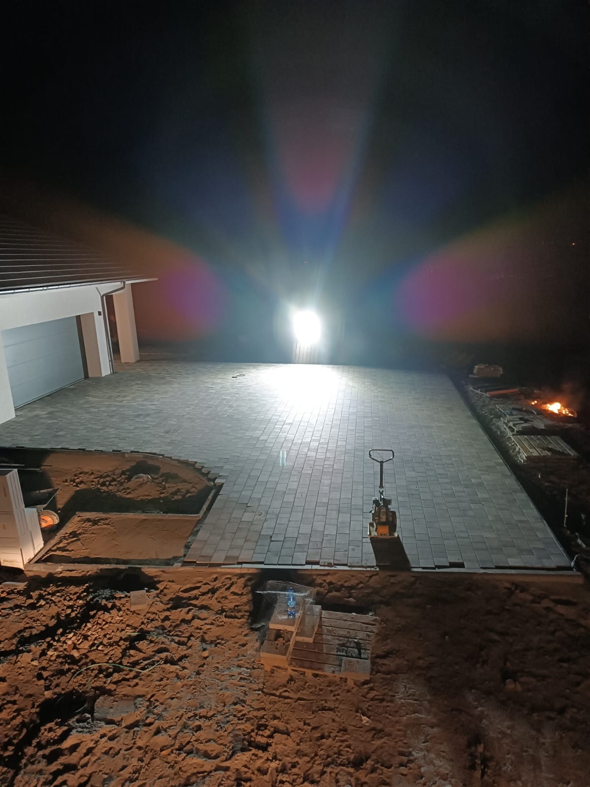 Nocna sceneria z nowo ułożoną kostką na podjeździe, widoczna zagęszczarka wibracyjna, niezakończone obrzeża z piaskiem i paleta z wodą, oświetlone mocnym światłem reflektora.