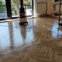 Błyszcząca, odnowiona podłoga parkietowa w jodełkę po zabiegu, z widocznym odkurzaczem przemysłowym i szlifierką podłogową, jasne wnętrze z dużymi oknami i schodami z drewnianą balustradą.