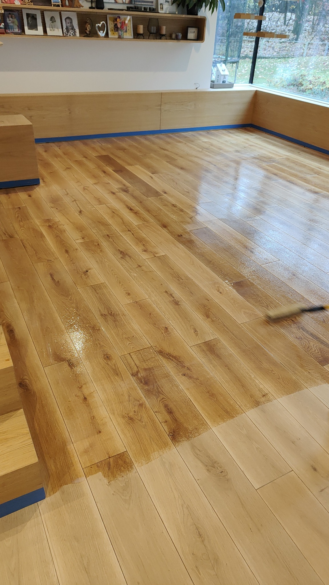 Aplikacja lakieru na parkiet za pomocą wałka, widoczne odświeżone i surowe deski, zabezpieczone taśmą ściany i meble.