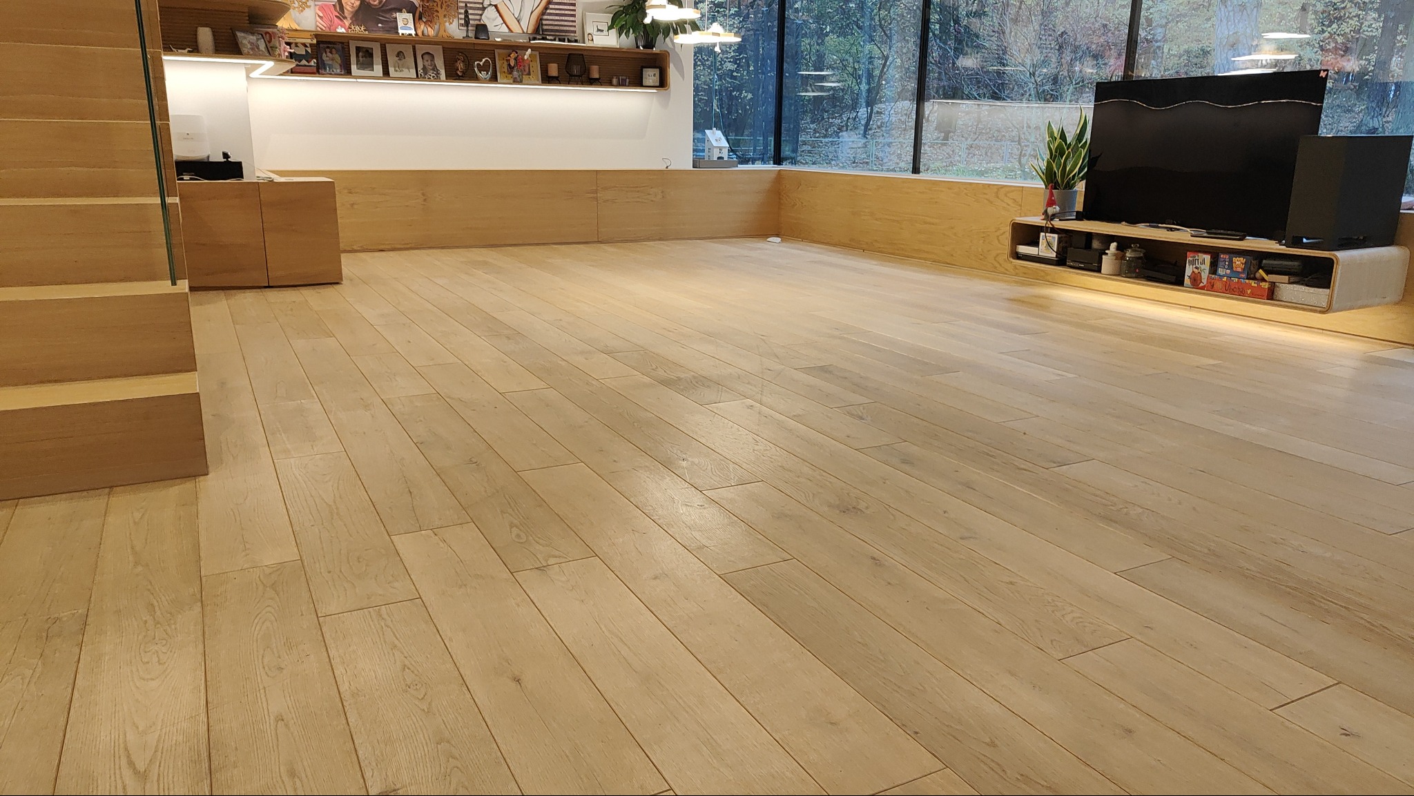 Jasny, drewniany parkiet ułożony w przestronnym salonie z widokiem na zewnątrz przez duże okno, schody z drewnianymi stopniami i szklaną balustradą po lewej stronie.