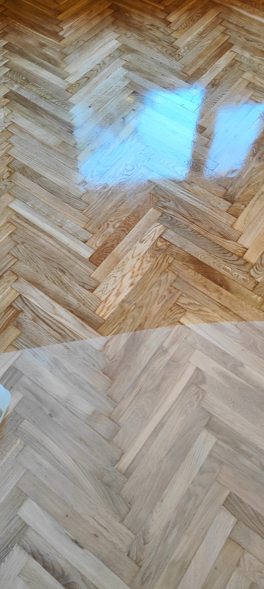 Fragment parkietu ułożonego we wzór jodełki, z widoczną różnicą w kolorze i połysku pomiędzy dwoma fragmentami podłogi, prawdopodobnie po cyklinowaniu i lakierowaniu.