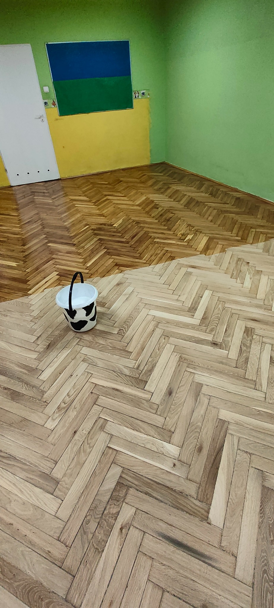 Częściowo cyklinowany parkiet w jodełkę, widoczny kontrast między odnowioną, ciemniejszą częścią a surowym, jaśniejszym drewnem; wiadro z motywem krowy stoi na nieobrobionej powierzchni, ściany...