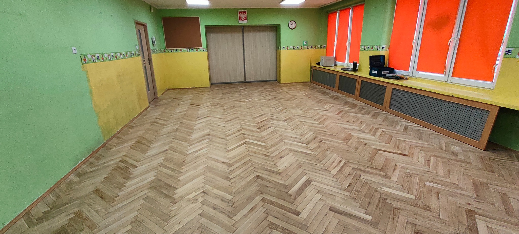 Parkiet ułożony we wzór jodełki w pustym pomieszczeniu z zielono-żółtymi ścianami i pomarańczowymi roletami okiennymi.