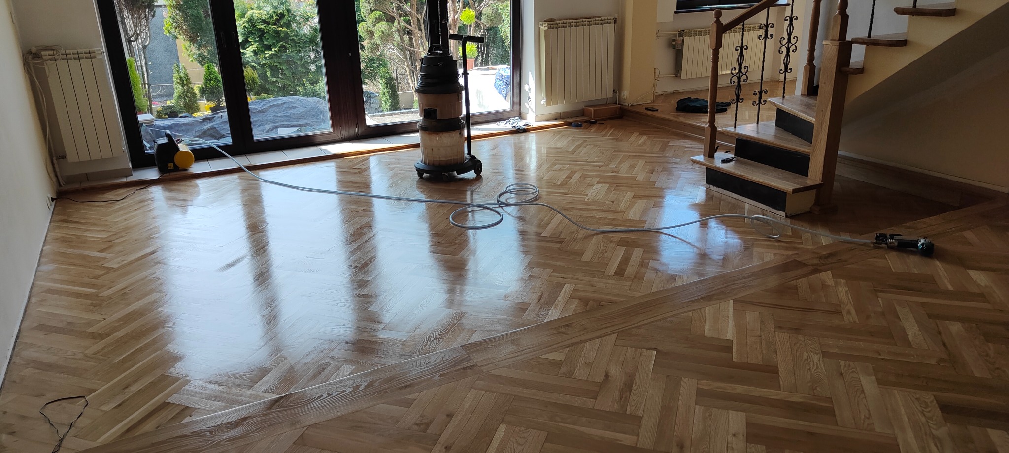 Błyszcząca, odnowiona podłoga parkietowa w jodełkę po zabiegu, z widocznym odkurzaczem przemysłowym i szlifierką podłogową, jasne wnętrze z dużymi oknami i schodami z drewnianą balustradą.