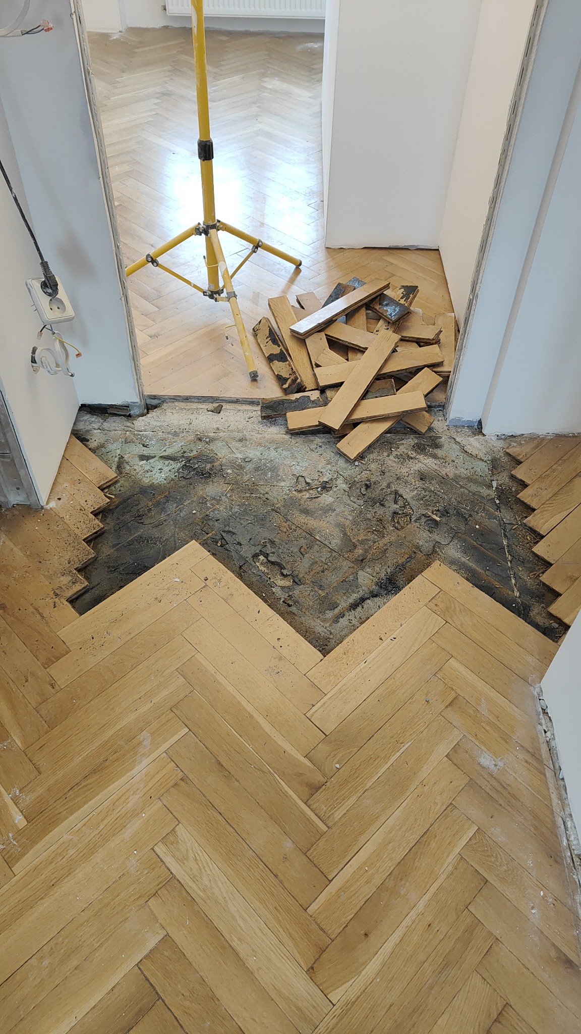 Fragment podłogi w jodełkę z częściowo usuniętym parkietem w Jaworznie, widoczny żółty statyw oświetleniowy i deski przygotowane do położenia.