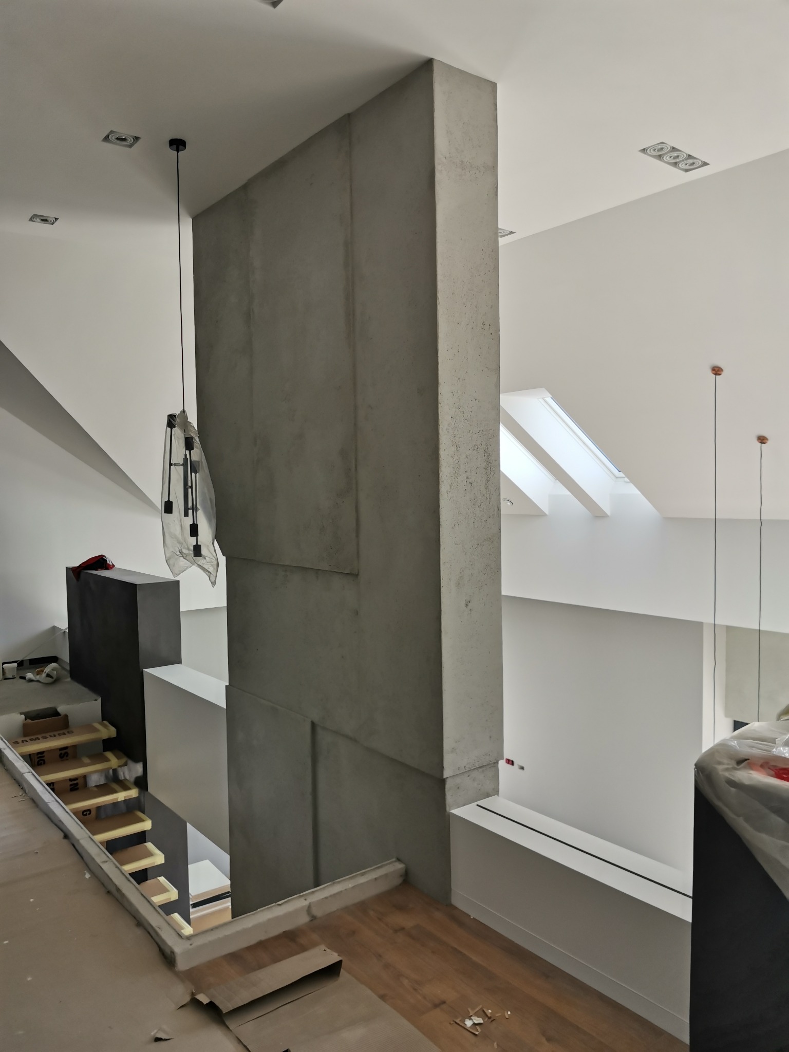 Wnętrze w trakcie wykańczania: betonowa kolumna z widocznymi łączeniami płyt, schody z zabezpieczeniami, nowoczesne oświetlenie wiszące i sufitowe, oraz okno dachowe w jasnym pomieszczeniu.