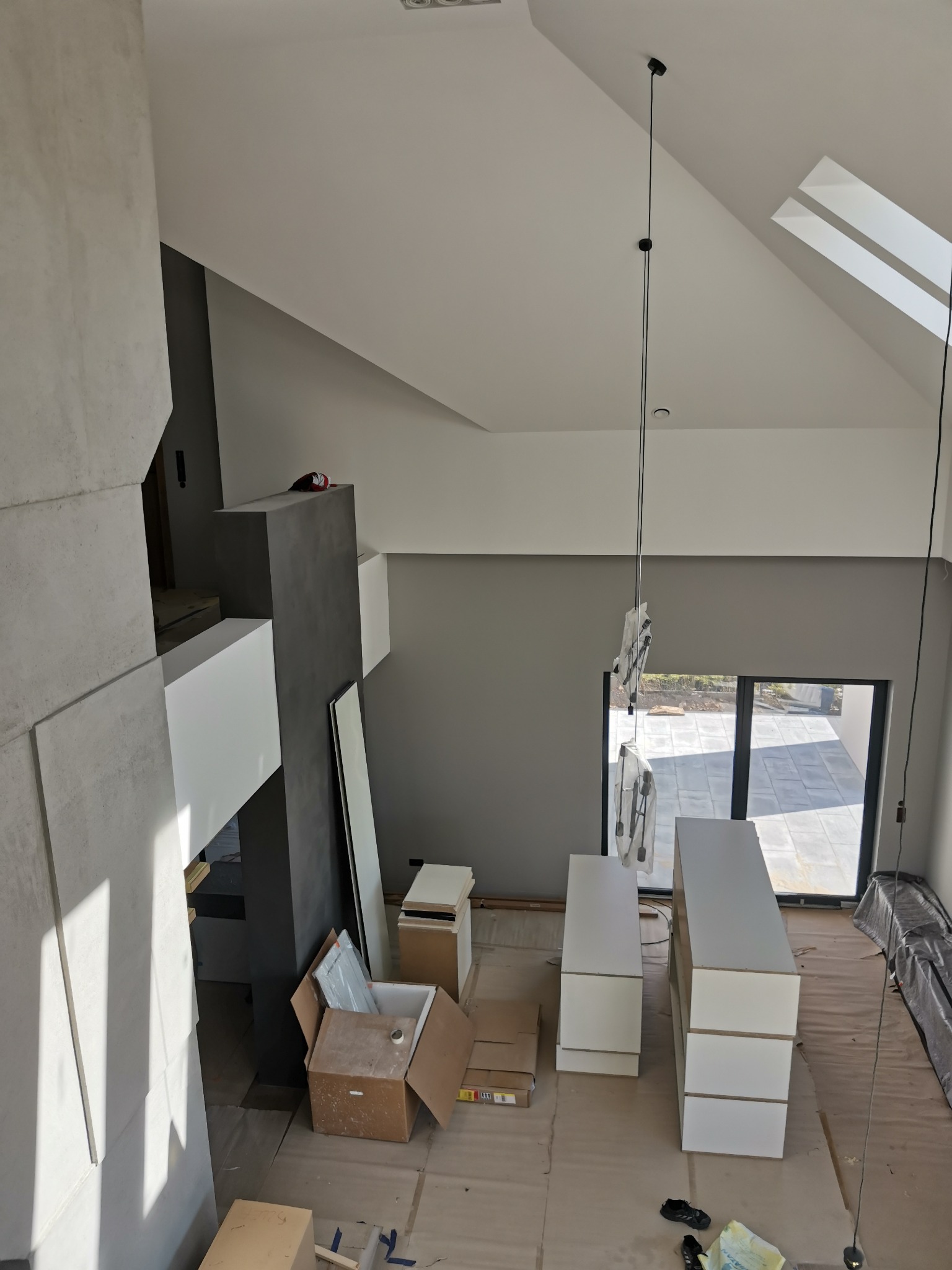 Wysoki, jasny pokój z surowymi ścianami, przygotowany do montażu mebli kuchennych, widok z góry na rozłożone kartony i zabezpieczoną podłogę.