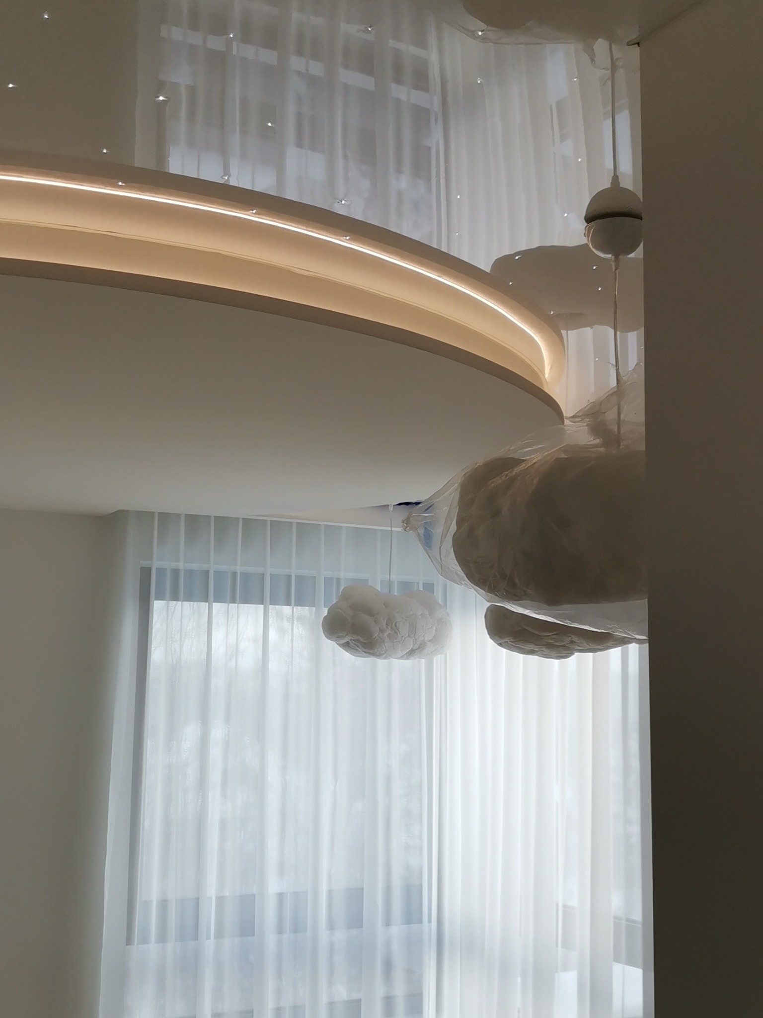 Nowoczesne oświetlenie sufitowe z podświetlanym okręgiem i dekoracyjnymi chmurkami zawieszonymi na żyłkach, widoczne okno z delikatnymi białymi zasłonami.