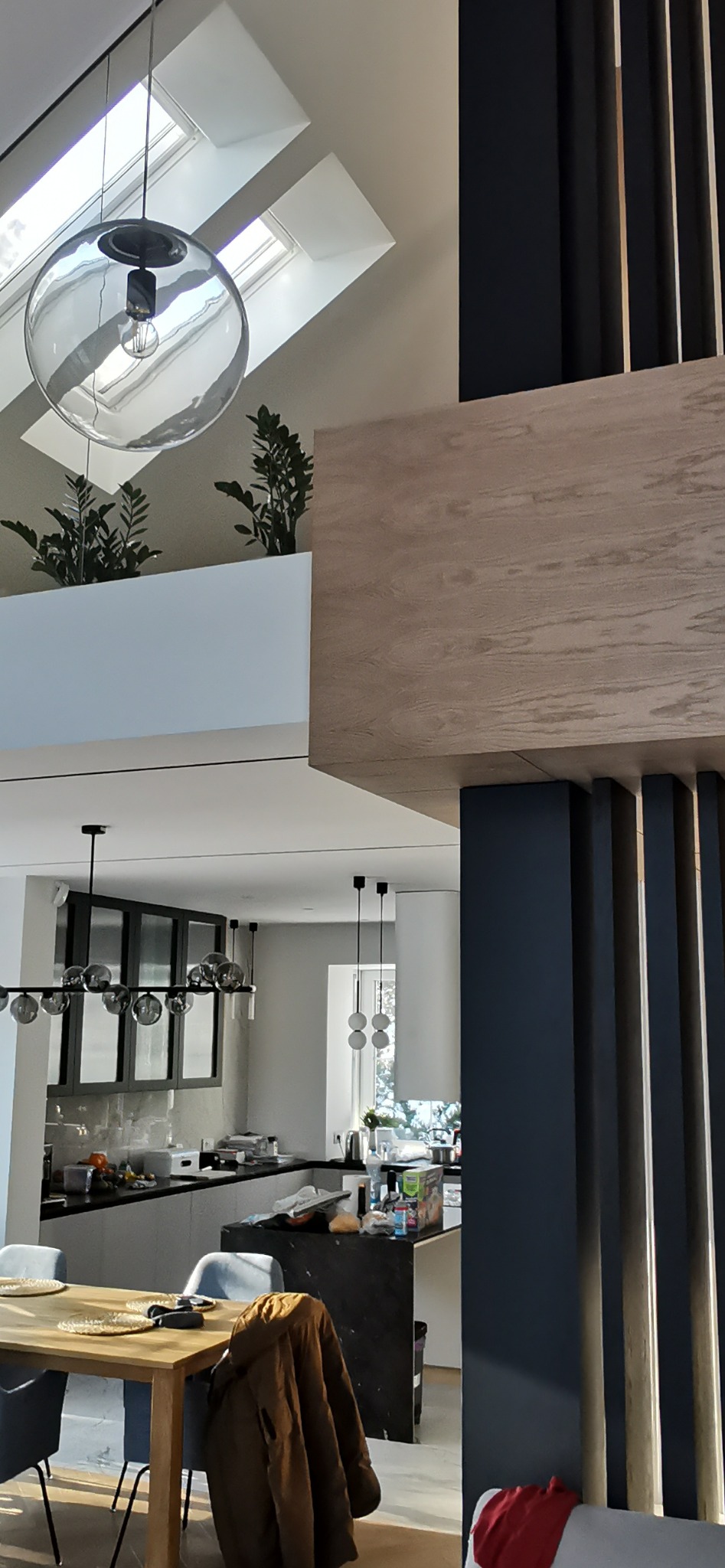 Wnętrze nowoczesnej kuchni z wyspą i jadalnią, widok na przeszklony sufit z roślinami, oświetlenie w postaci szklanej kuli i lamp wiszących.