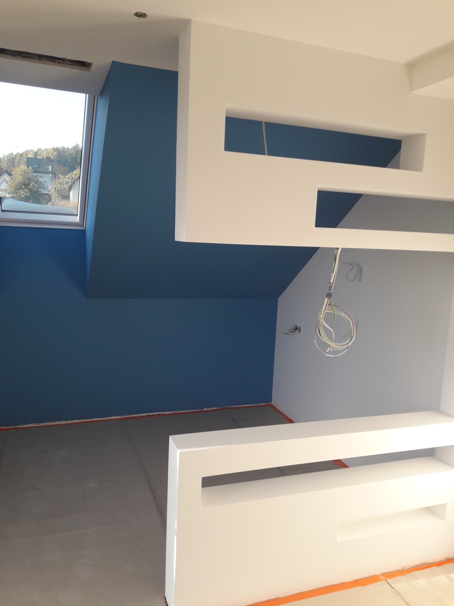 Poddasze z pomalowaną na niebiesko ścianą i elementami dekoracyjnymi z płyt gipsowo-kartonowych, widoczne wystające przewody elektryczne.