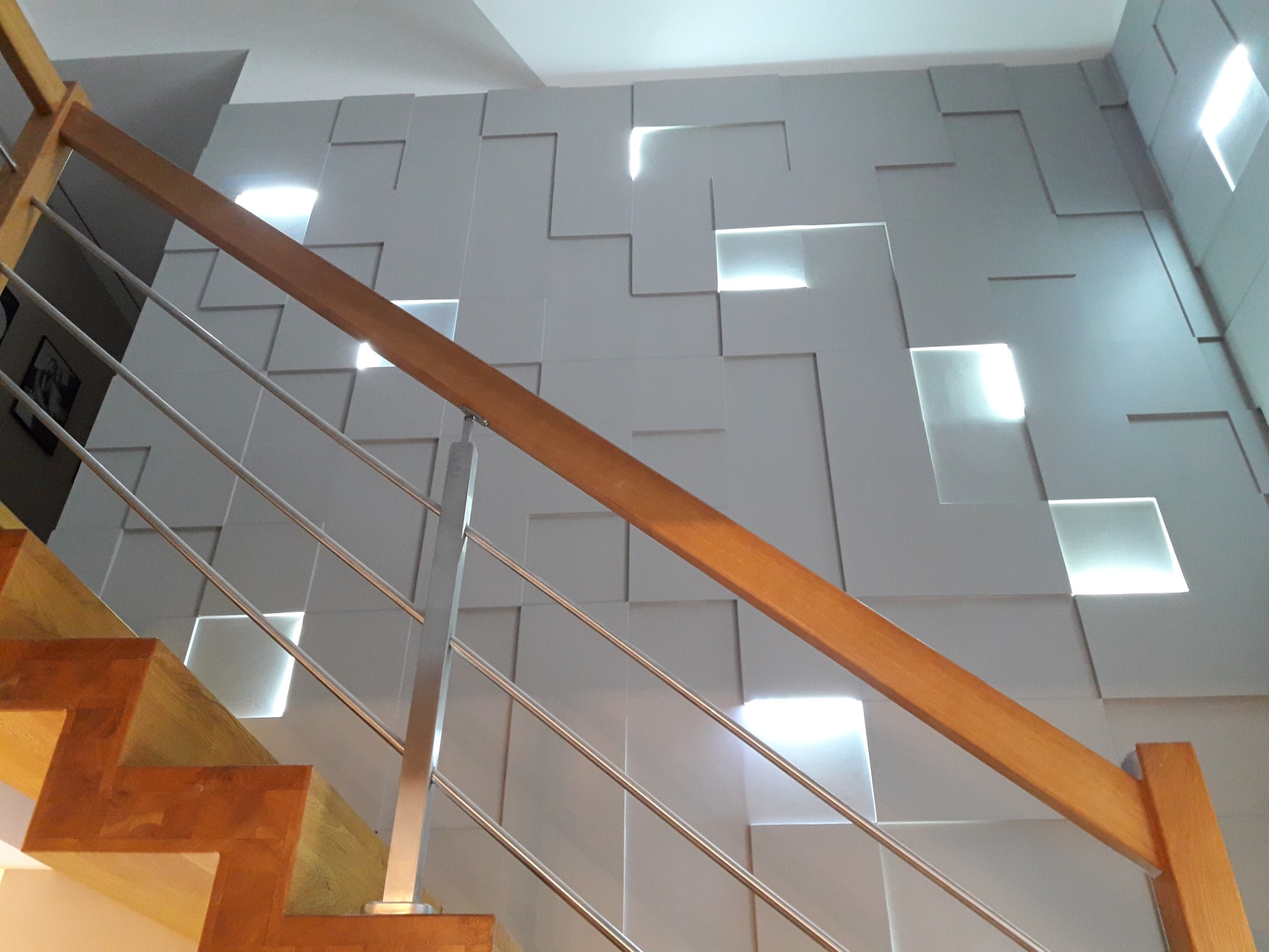 Nowoczesna ściana z paneli 3D oświetlona od tyłu, widok z dołu schodów z drewnianymi stopniami i metalową balustradą.