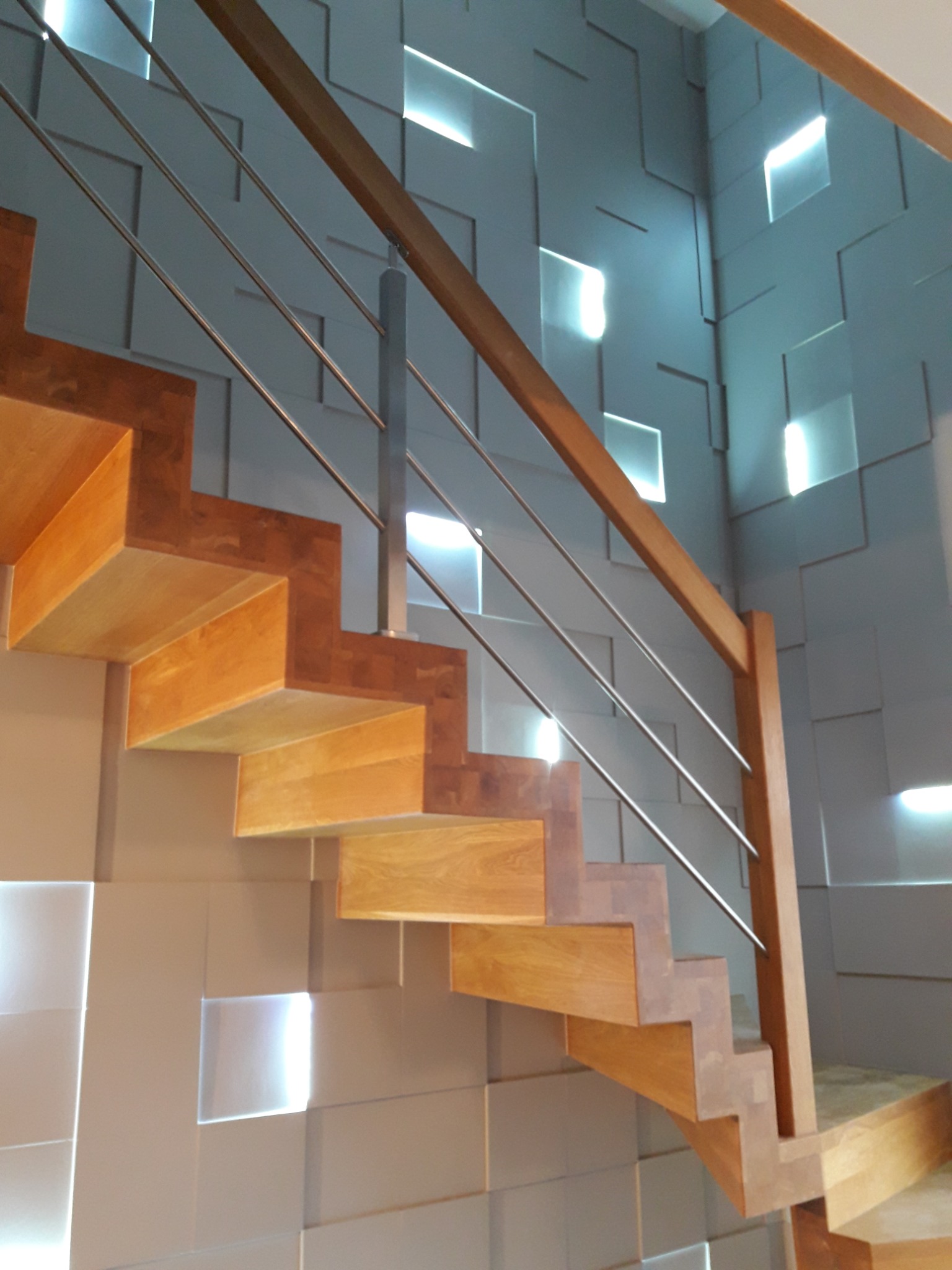 Drewniane schody z metalową balustradą na tle ściany z kwadratowych paneli z efektem świetlnym.