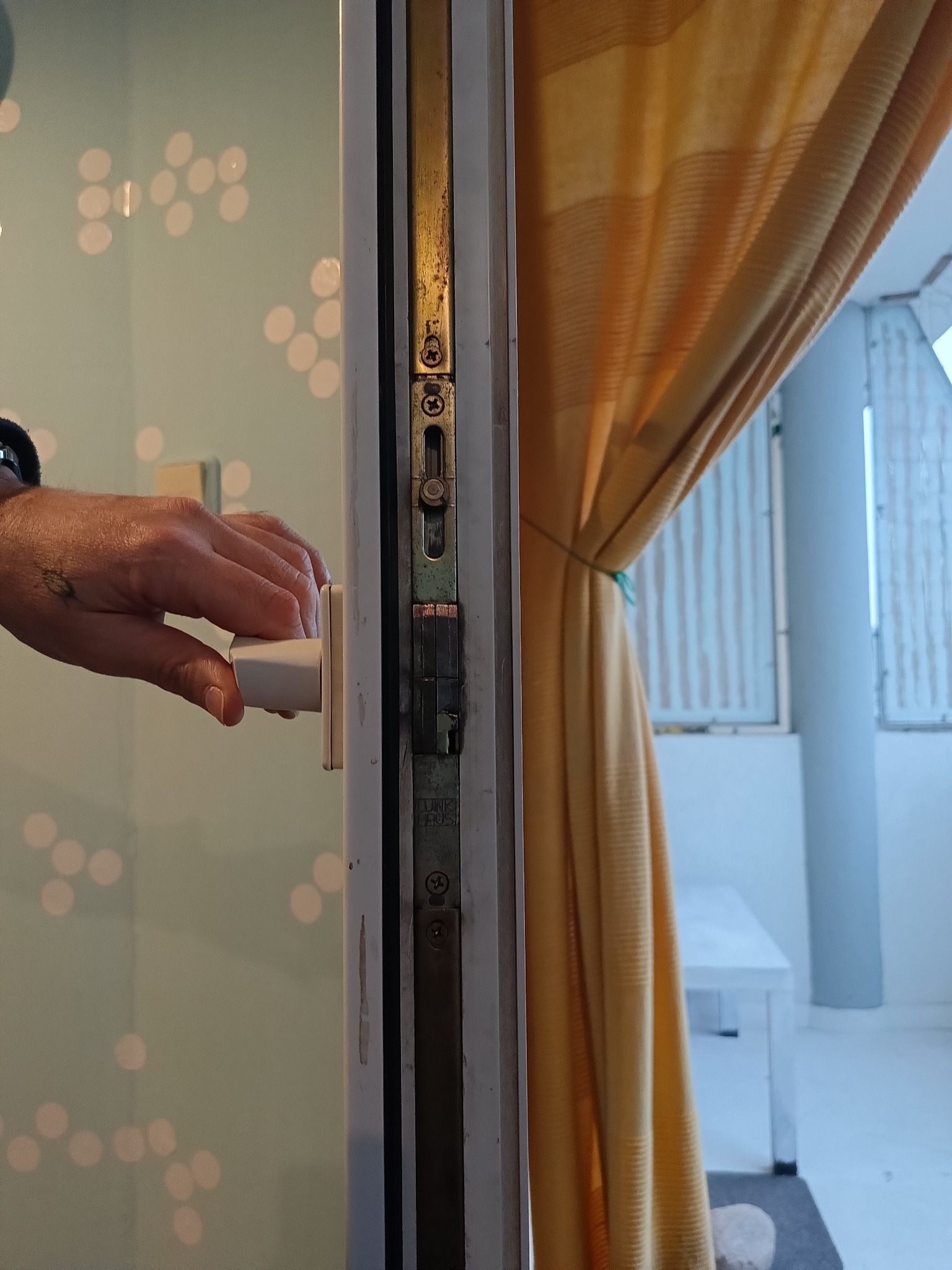 Dłoń z tatuażem obraca białą klamkę okna balkonowego; widoczne ślady użytkowania na metalowej ramie okna, w tle żółta zasłona i jasne wnętrze balkonu.