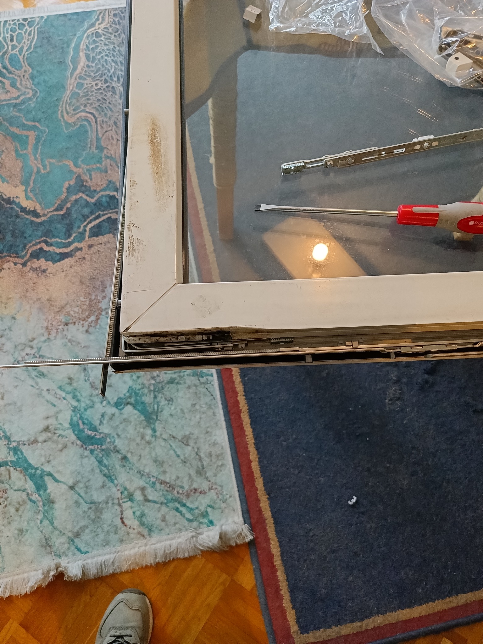 Detal naprawy okna: widoczny mechanizm okienny, śrubokręt z czerwoną rączką, okno z widocznymi śladami użytkowania, fragment dywanu z wzorem w odcieniach błękitu i fragment dywanu w kolorze...