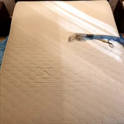 Czyszczenie materaca beżowym odkurzaczem piorącym. Widoczny fragment wypranej powierzchni i urządzenie z wężem. Dwuosobowy materac na łóżku.