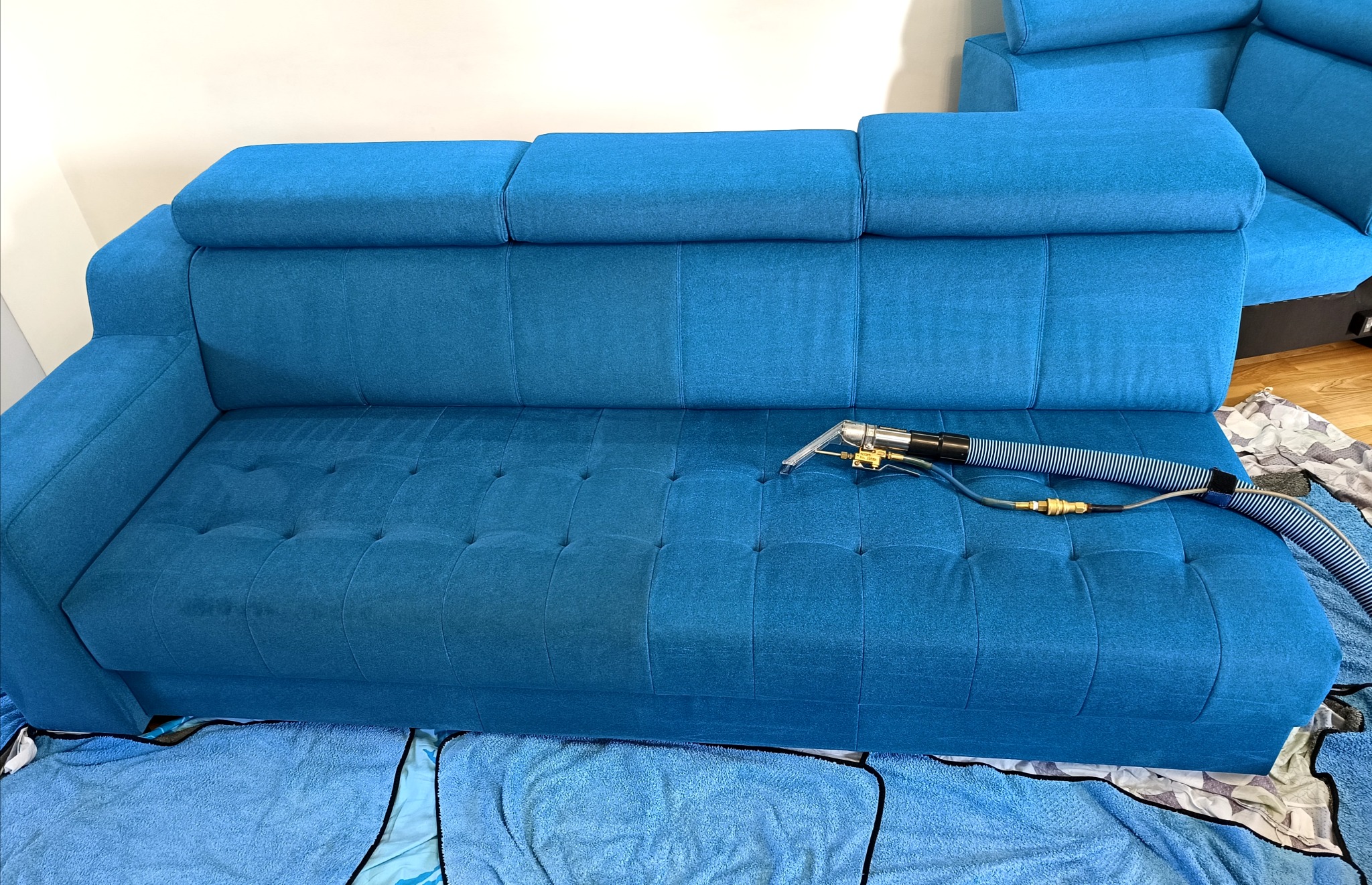 Turkusowa kanapa z widocznym efektem prania: połowa czysta, połowa z plamami. Dysza odkurzacza piorącego na powierzchni. Ręcznik zabezpieczający pod spodem.