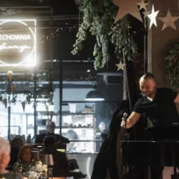 Występujący mężczyzna z mikrofonem na scenie restauracji, dekoracje z zielonych gałązek i neon 'Cechownia'