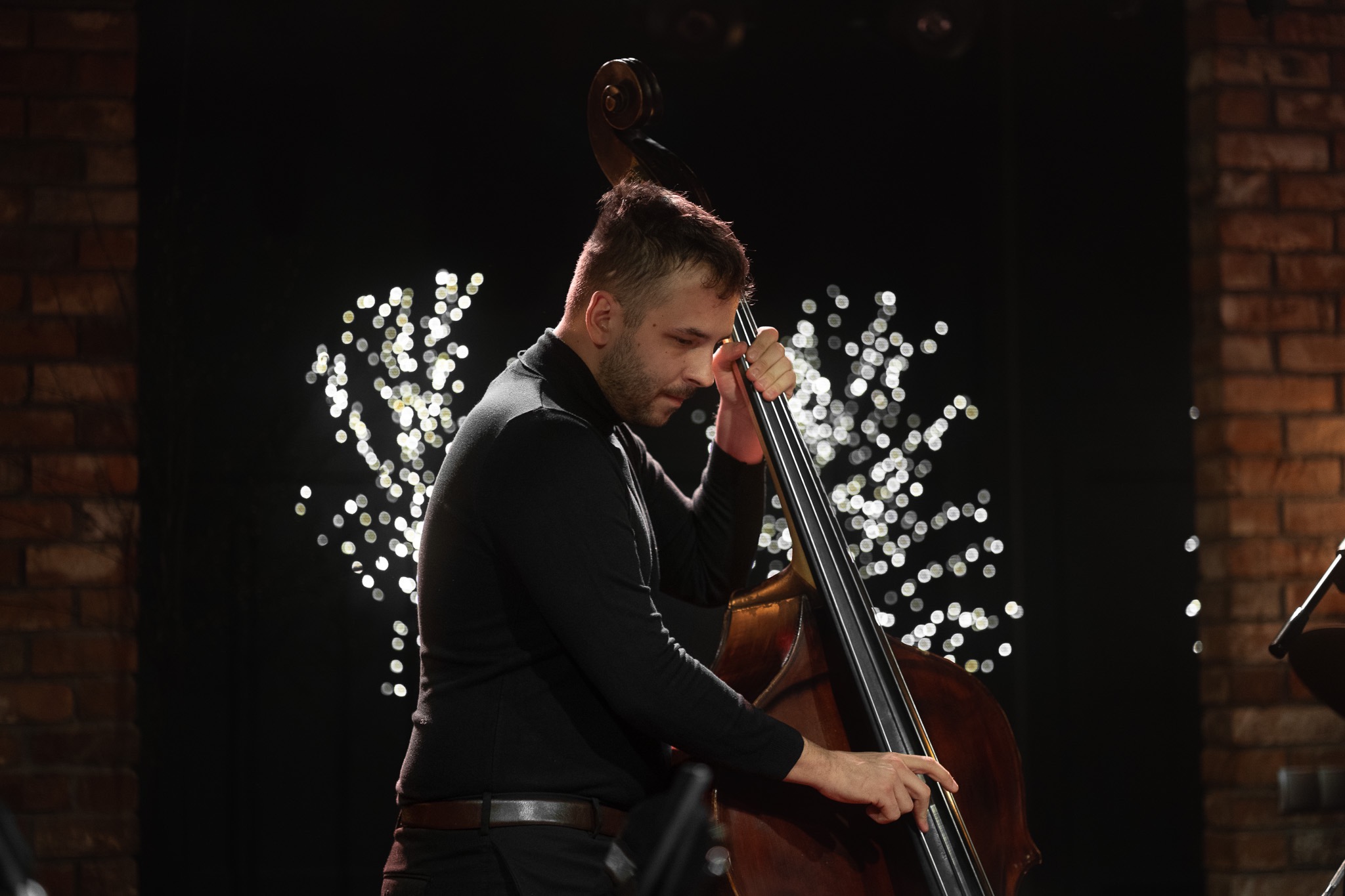 Mężczyzna w czarnym golfie gra na kontrabasie na tle ceglanej ściany i światełek.