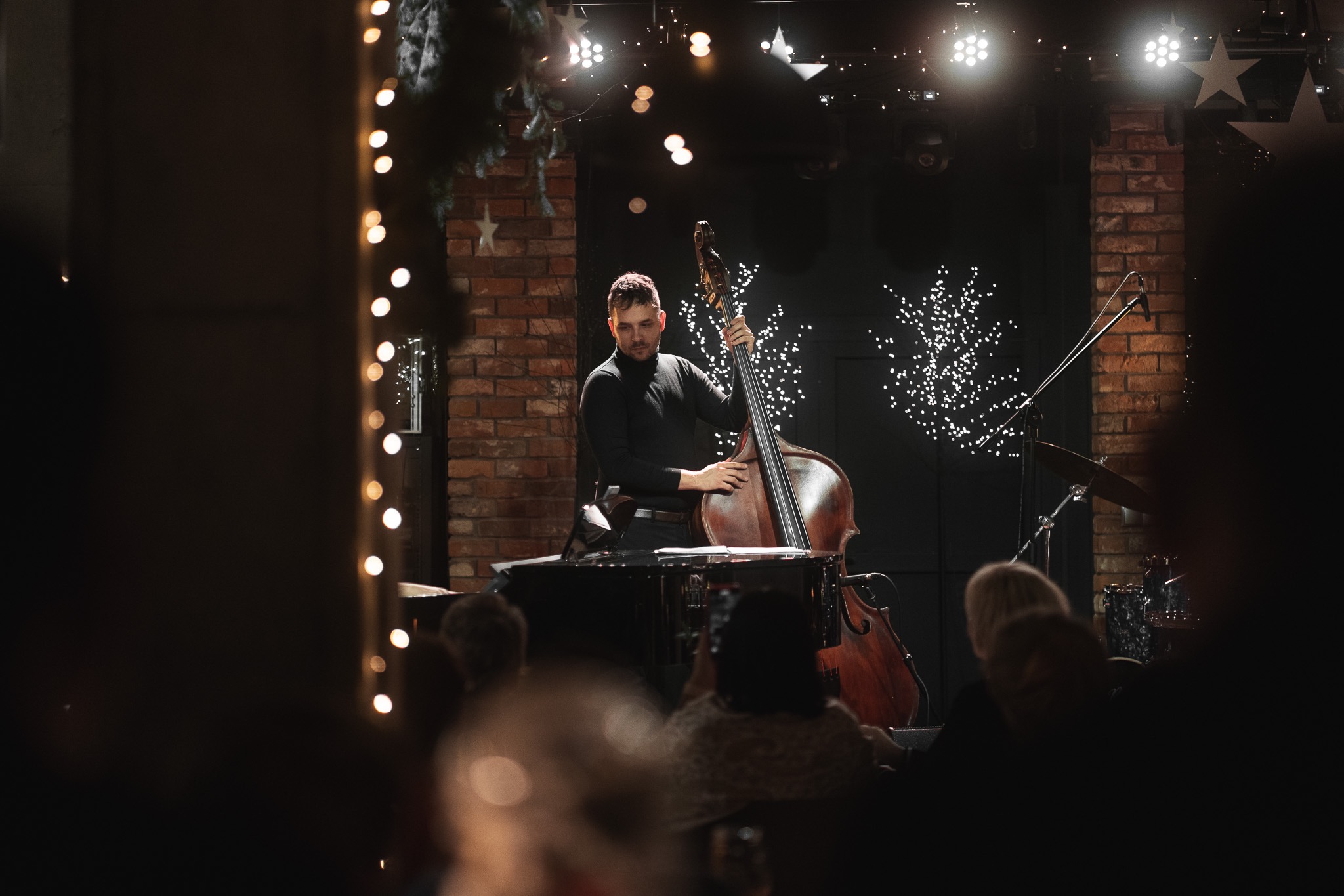 Kontrabasista w czarnym golfie na scenie z ceglanym filarem i dekoracjami świetlnymi, widoczny z perspektywy widowni.
