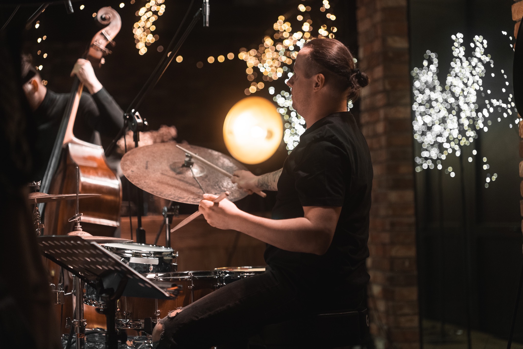Muzyk grający na perkusji podczas występu na żywo, w tle kontrabasista i dekoracyjne światła.