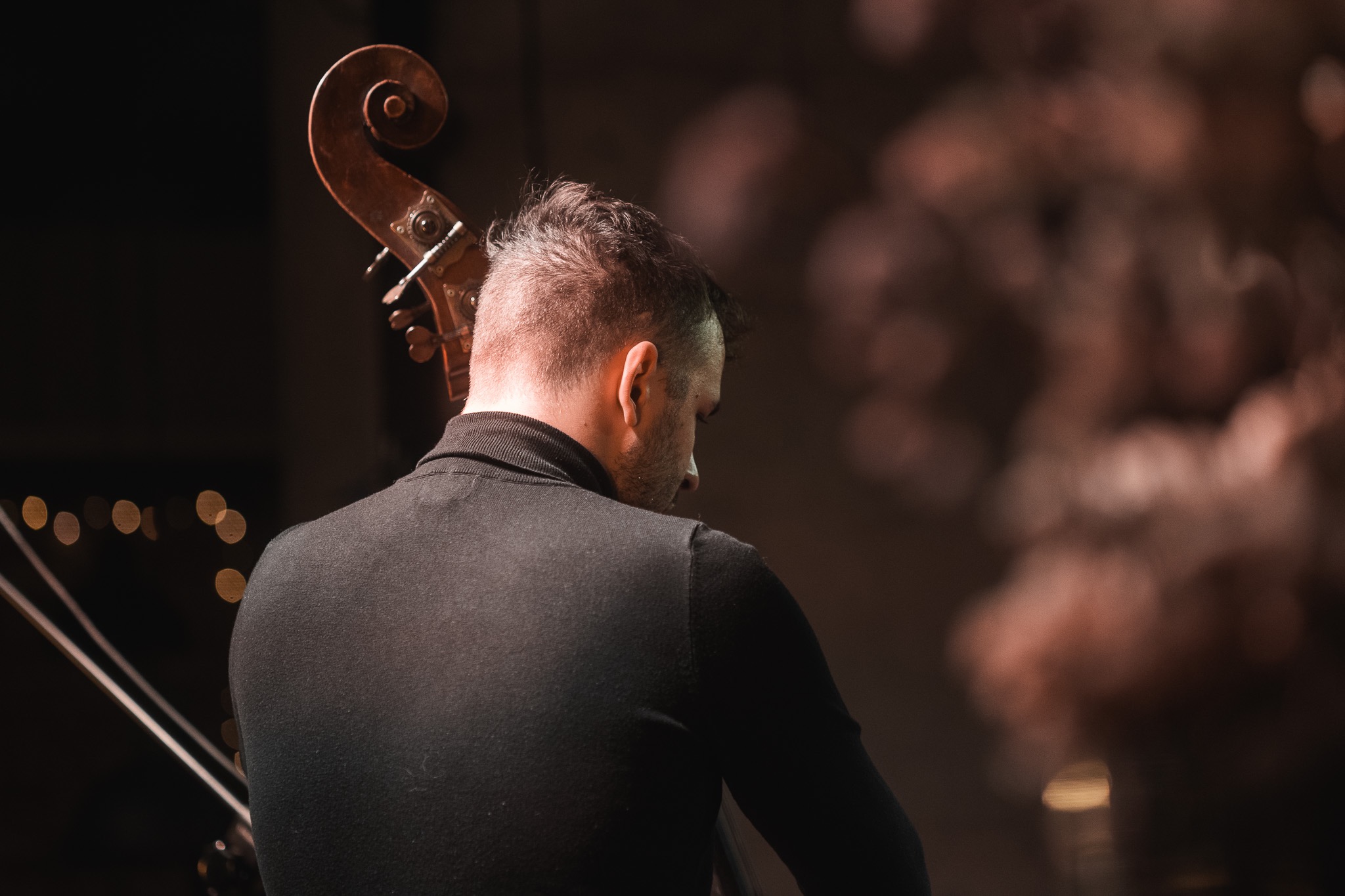 Mężczyzna w czarnym golfie gra na kontrabasie, widok z tyłu, z rozmyciem w tle.