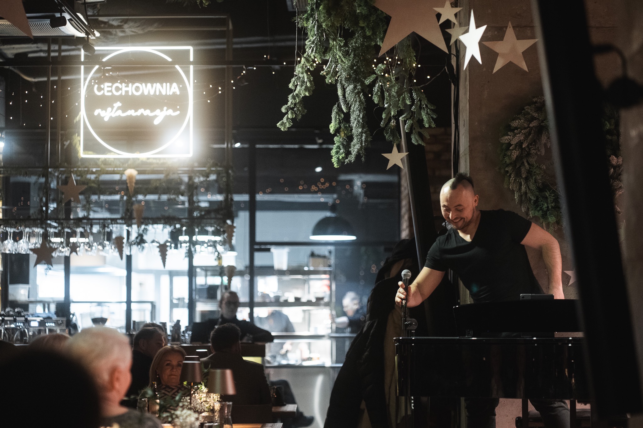 Występujący mężczyzna z mikrofonem na scenie restauracji, dekoracje z zielonych gałązek i neon 'Cechownia'