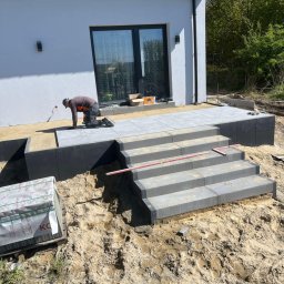 M-Bruk - Pracownik w trakcie układania szarej kostki brukowej na schodach i tarasie przed nowoczesnym domem z białymi ścianami i dużym oknem tarasowym, widoczne palety z kostką i poziomica.