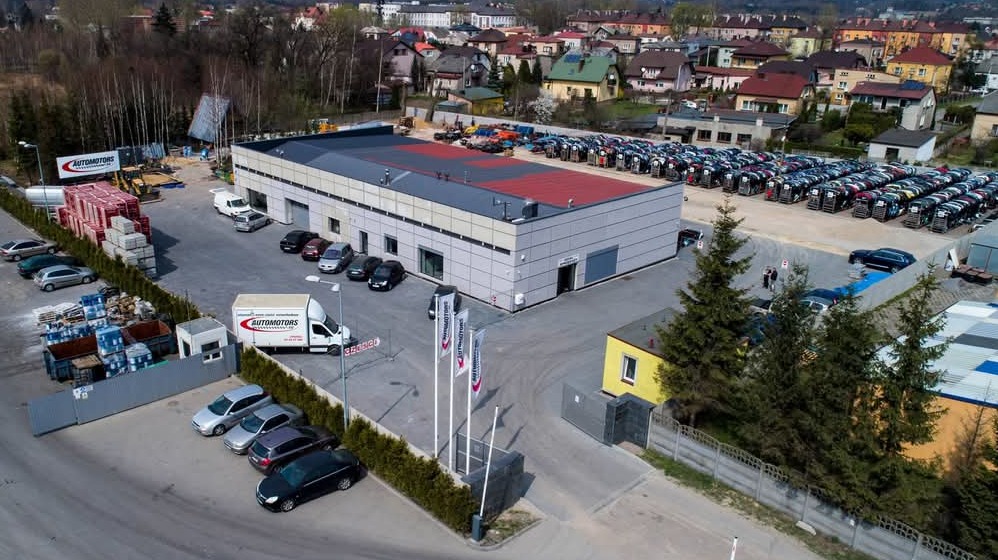 Widok z góry na plac firmy Automotors z budynkiem biurowym, zaparkowanymi samochodami, stertą kostki brukowej i banerem reklamowym.