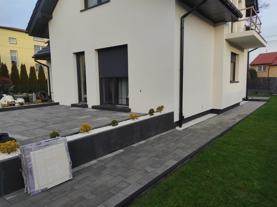 Nowoczesny taras i ścieżka z szarej kostki brukowej, obrzeża z czarnego betonu, białe kamyczki dekoracyjne wzdłuż ściany budynku, fragment trawnika i domu w tle.