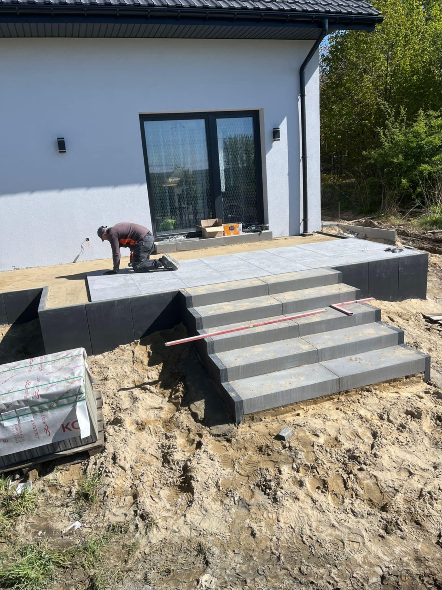Pracownik w trakcie układania szarej kostki brukowej na schodach i tarasie przed nowoczesnym domem z białymi ścianami i dużym oknem tarasowym, widoczne palety z kostką i poziomica.