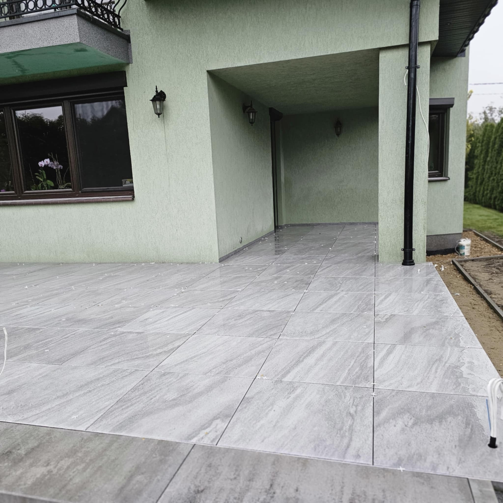 Jasnoszare płytki imitujące kamień, ułożone na tarasie przed domem z zieloną elewacją, widoczne krzyżyki dystansowe.