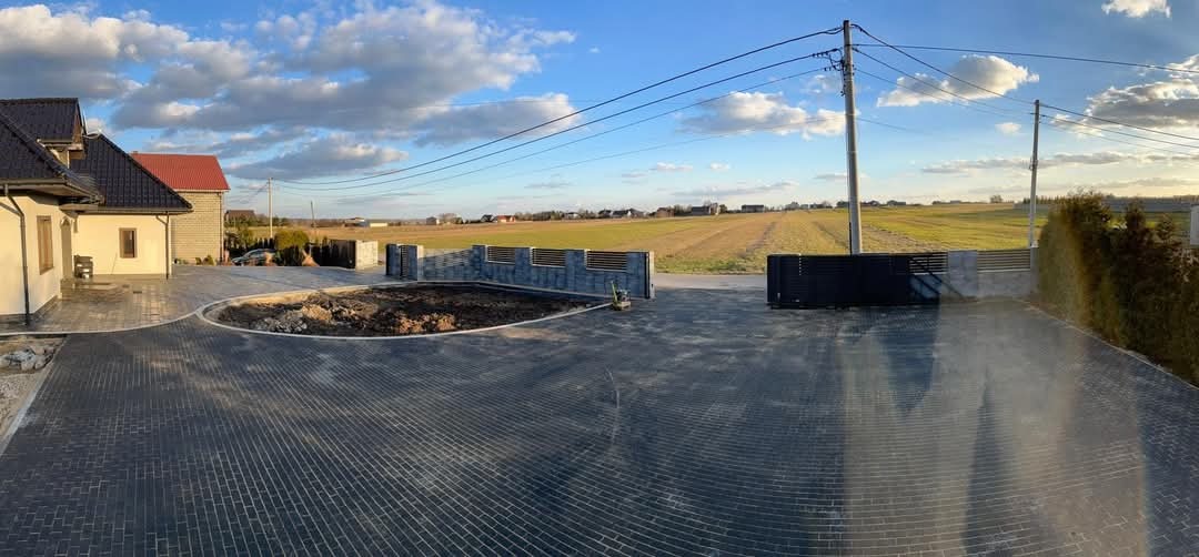 Ułożona ciemnoszara kostka brukowa na podjeździe domu z przygotowanym miejscem na trawnik, widoczne słupki ogrodzeniowe i brama wjazdowa, w tle pole i linia wysokiego napięcia.