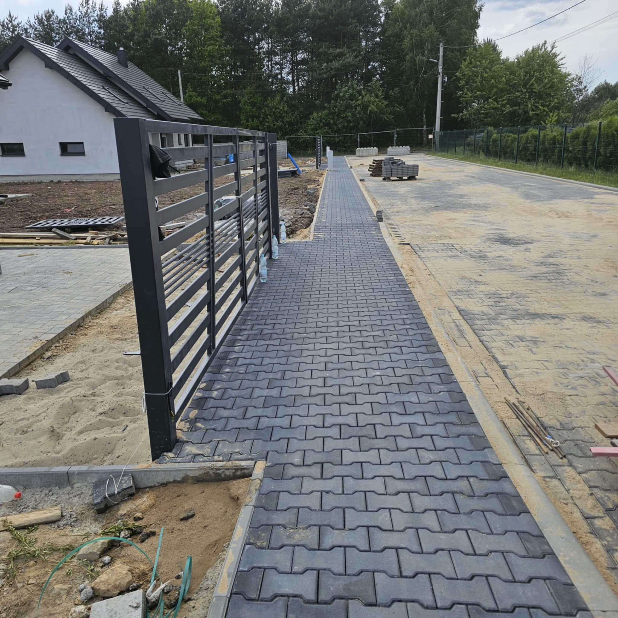 Nowo ułożona, szara kostka brukowa w kształcie litery H prowadząca wzdłuż ogrodzenia posesji, z widocznymi elementami piasku i przygotowanymi kostkami w tle.