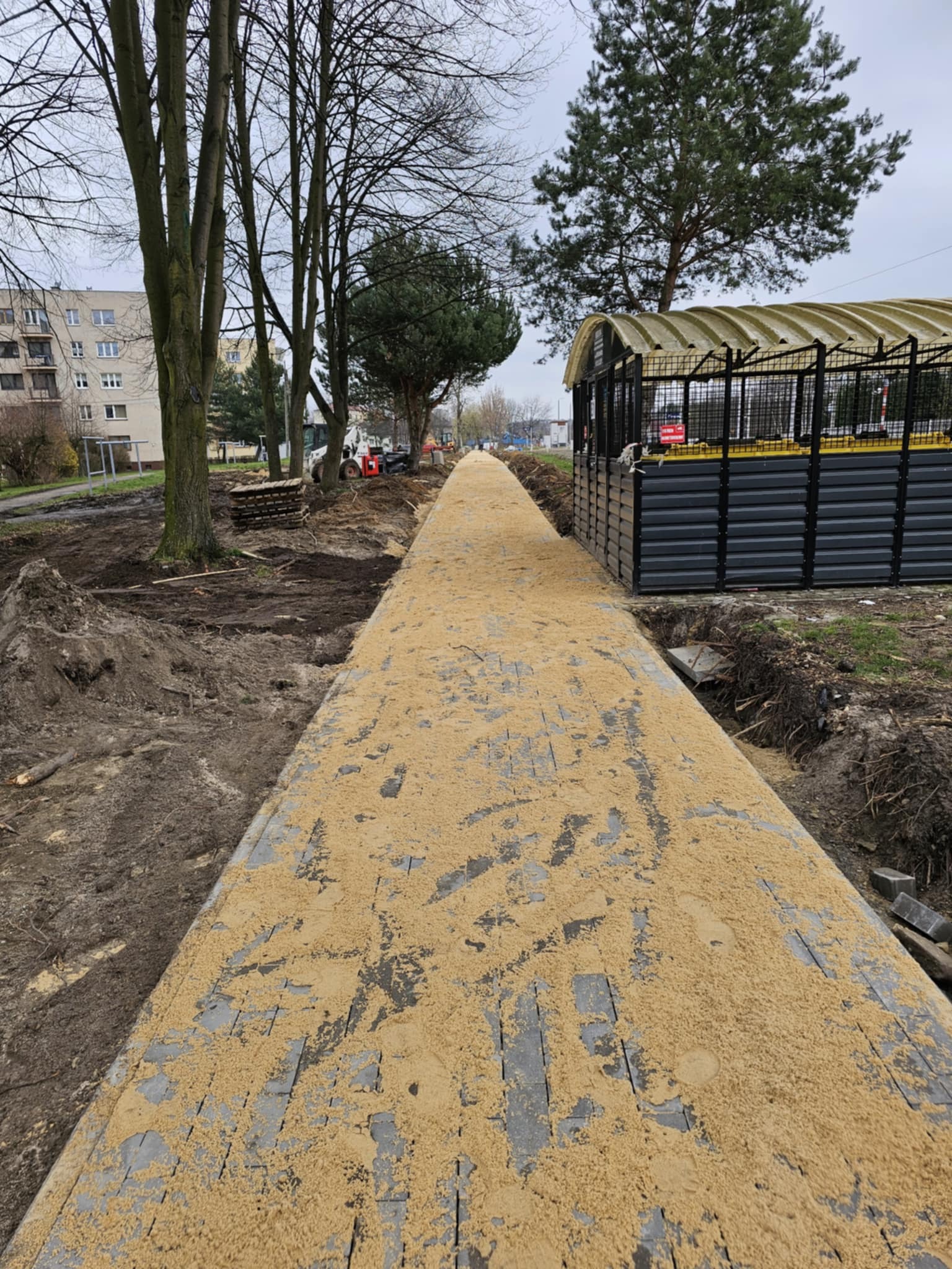 Długa ścieżka z kostki brukowej, zasypana piaskiem, w trakcie budowy w parku miejskim, z widocznymi maszynami budowlanymi w tle i drzewami po bokach.