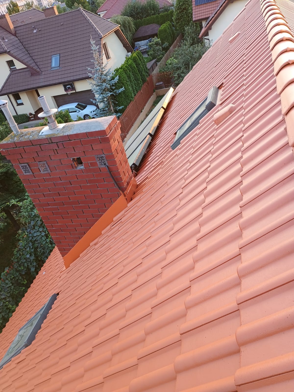 Czerwona dachówka ceramiczna na skośnym dachu z oknem dachowym i ceglanym kominem z kratkami wentylacyjnymi, widok z góry na tle domów i zieleni.