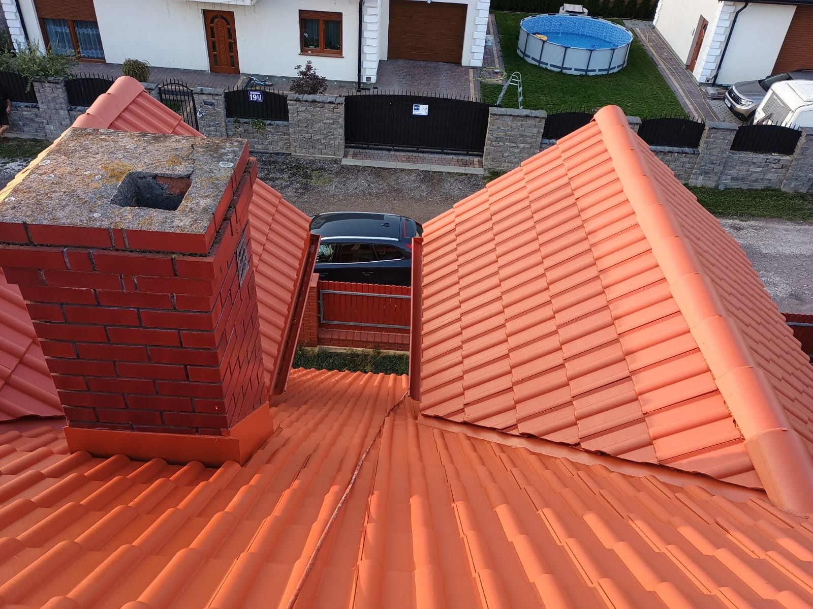 Czerwony dach z dachówki ceramicznej widziany z góry, z kominem z cegły i widokiem na posesję z samochodem i basenem w tle.