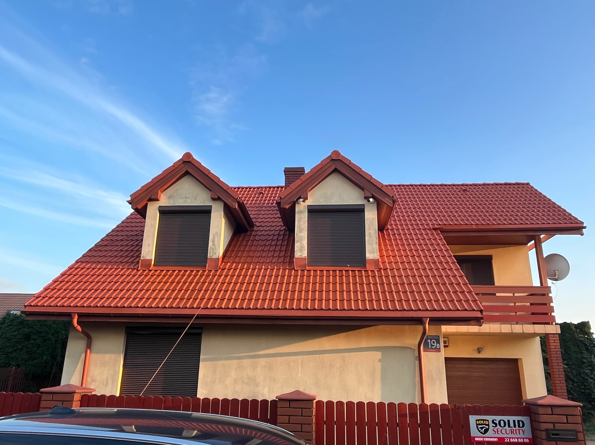 Dom jednorodzinny z czerwoną dachówką i ciemnymi roletami zewnętrznymi, widoczny numer domu 19B oraz fragment ogrodzenia i samochodu.