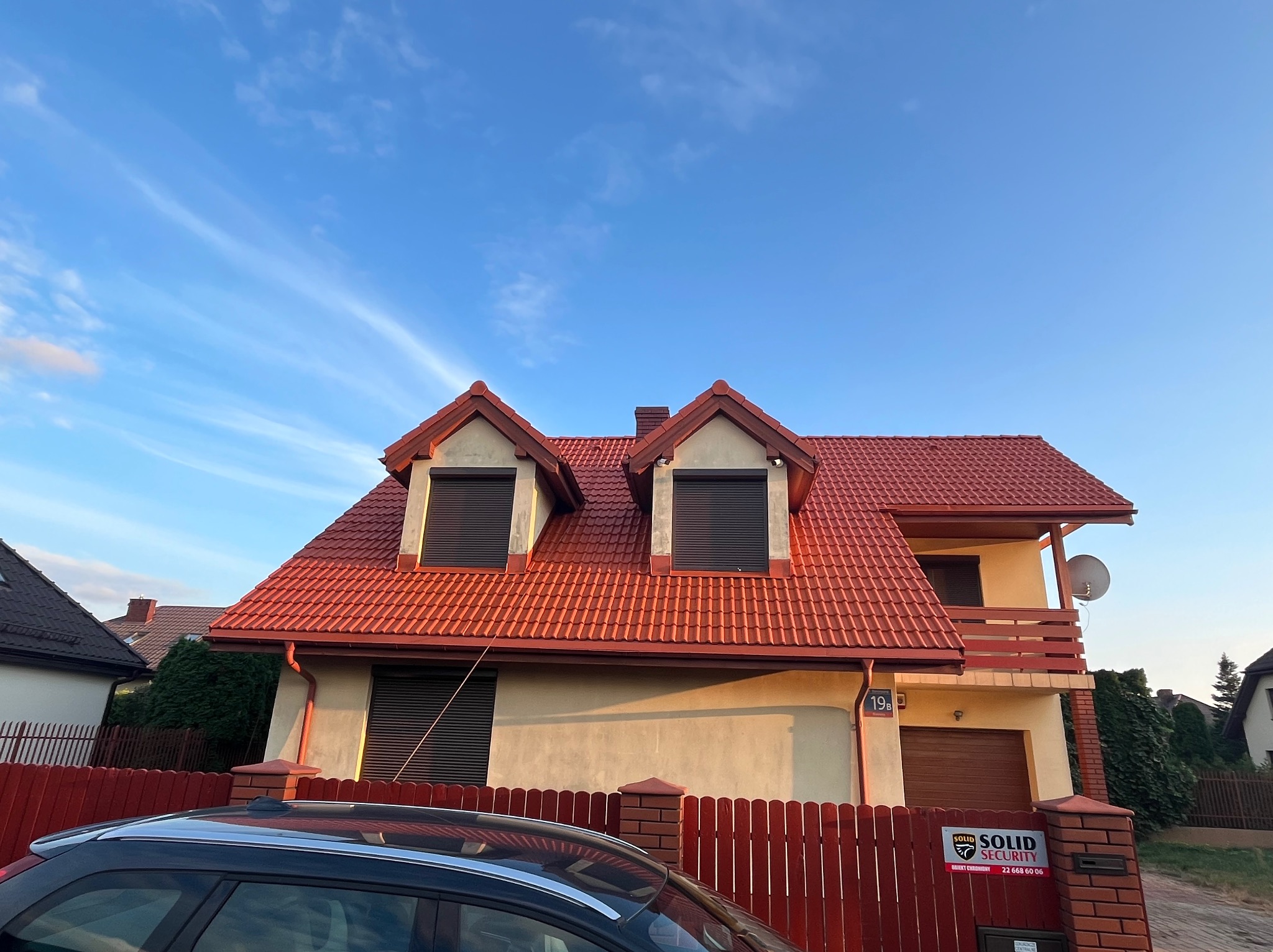 Dwupiętrowy dom z czerwoną dachówką i drewnianym ogrodzeniem, widoczny fragment samochodu na pierwszym planie, błękitne niebo w tle.