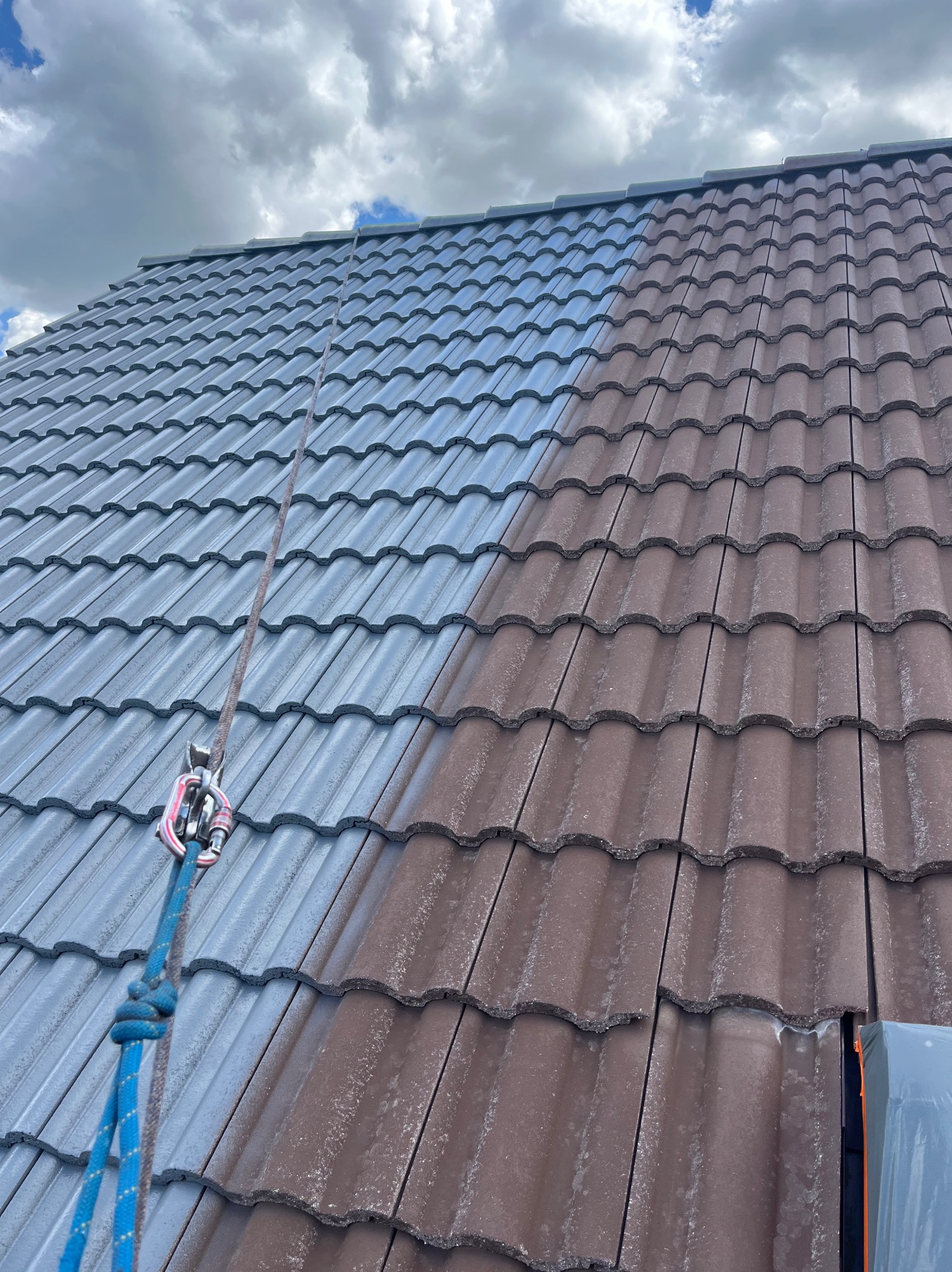 Częściowo umyty dach z dachówki ceramicznej, widoczna różnica między czystą, szarą powierzchnią a brudną, brązową; lina zabezpieczająca z karabińczykiem. Ujęcie z dołu, pochmurne niebo w tle.