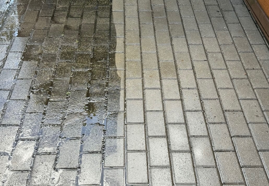 Częściowo umyta kostka brukowa, widoczna różnica w kolorze i czystości między umytą a nieumytą powierzchnią, mokra nawierzchnia.