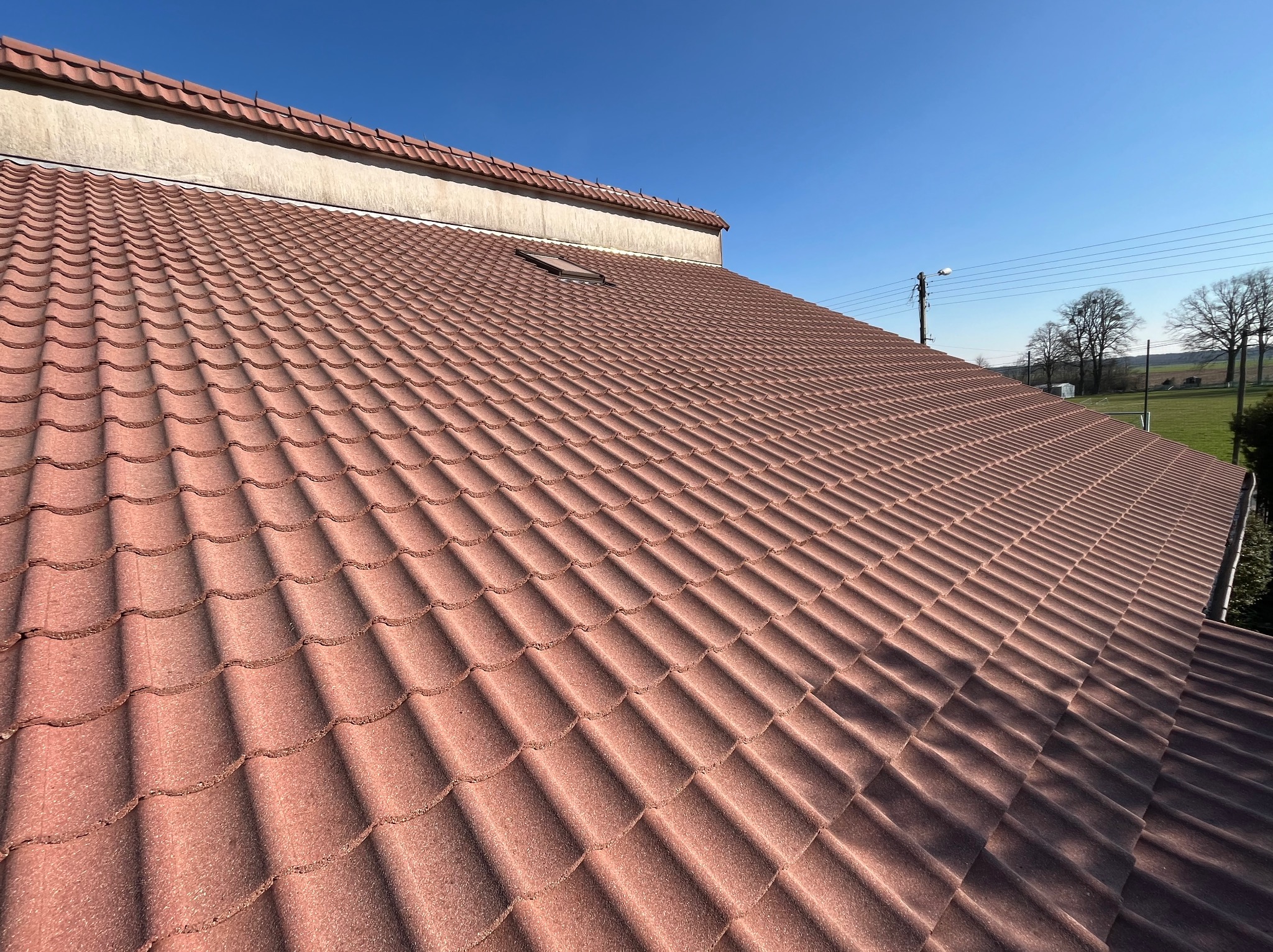 Czerwony dach z dachówki ceramicznej, widok z góry, z oknem dachowym, na tle błękitnego nieba i zielonego krajobrazu.