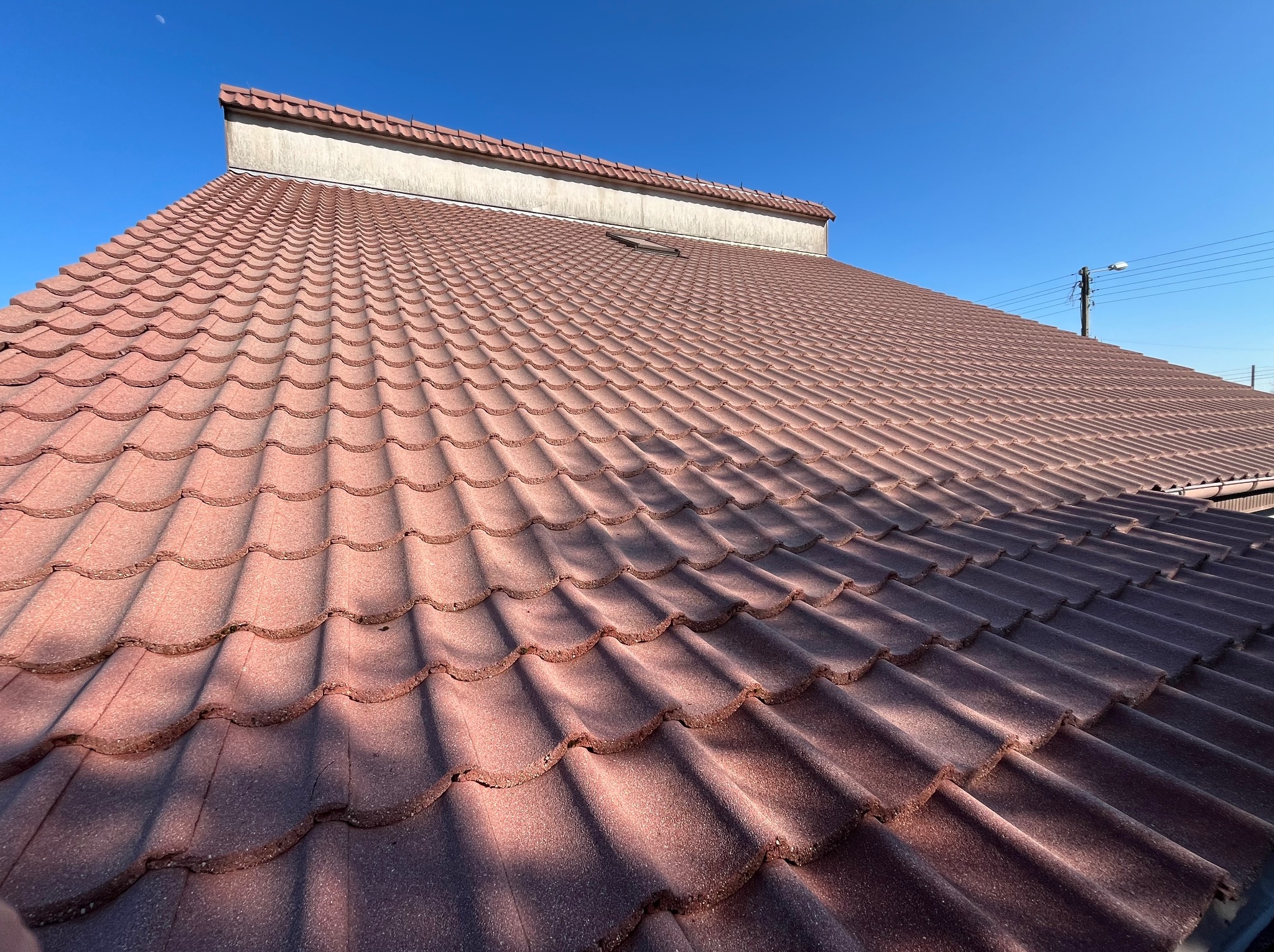 Czerwono-brązowy dach z dachówki ceramicznej w kształcie fali, sfotografowany pod kątem, z widocznym fragmentem elewacji i słupem elektrycznym na tle błękitnego nieba.
