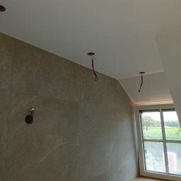 Robert BARTNIK - Wnętrze w trakcie prac wykończeniowych: ściana wyłożona płytkami imitującymi marmur, sufit z otworami na oświetlenie, widoczne przewody elektryczne, okno z widokiem na zielony krajobraz.