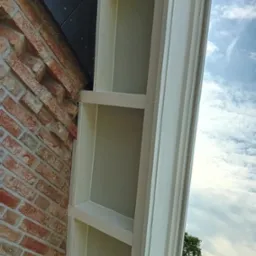 Fragment elewacji budynku z cegły klinkierowej, z widocznym białym, drewnianym elementem dekoracyjnym przypominającym pilaster, na tle błękitnego nieba z chmurami.