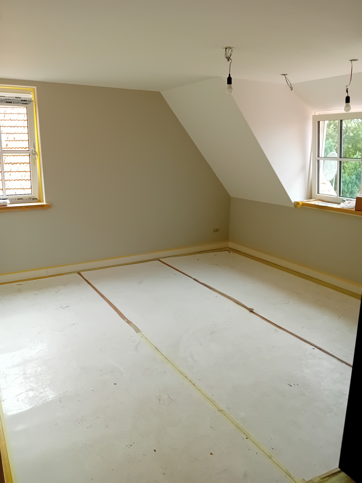 Pokój na poddaszu w trakcie remontu: ściany pomalowane na szaro, podłoga pokryta wylewką samopoziomującą, okna zabezpieczone taśmą malarską, widoczne wiszące żarówki.