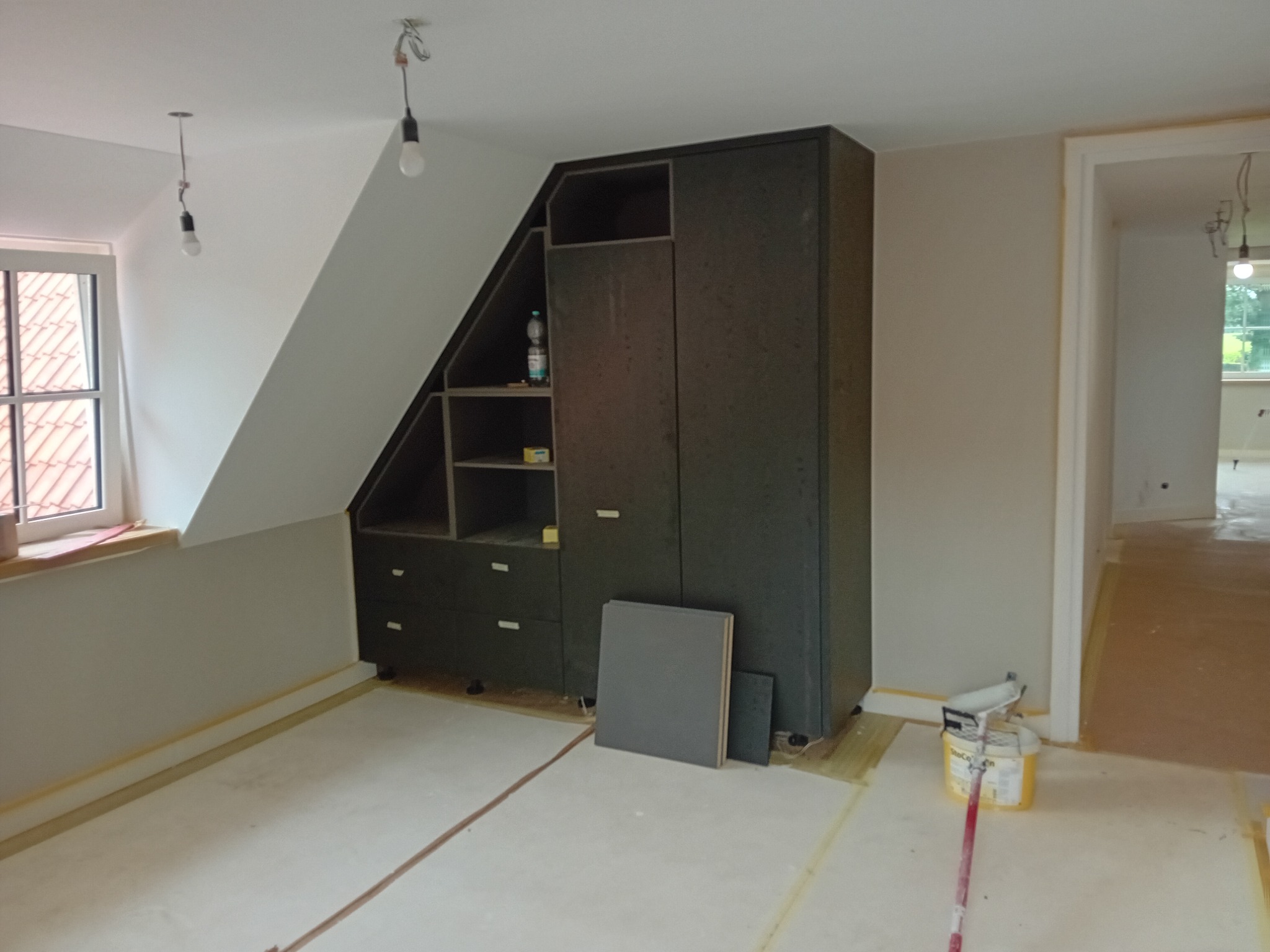 Wnętrze poddasza z zabudową szafy wnękowej z ciemnej płyty, widoczne niedokończone malowanie ścian, zabezpieczone taśmą malarską podłogi i wiadro z farbą.