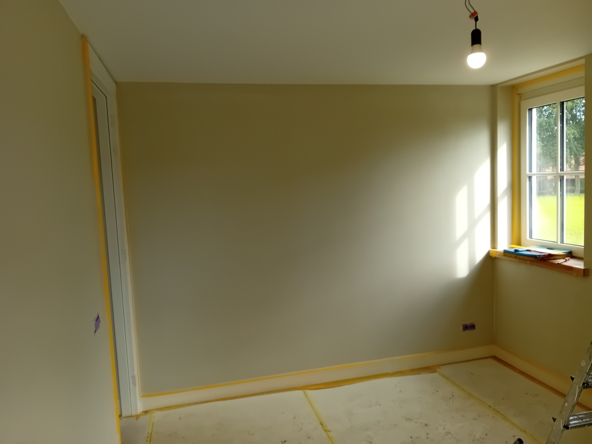 Pokój przygotowany do malowania: ściany zabezpieczone taśmą malarską, podłoga zakryta folią ochronną, widoczne gniazdko elektryczne i okno z parapetem, na którym leżą narzędzia malarskie.