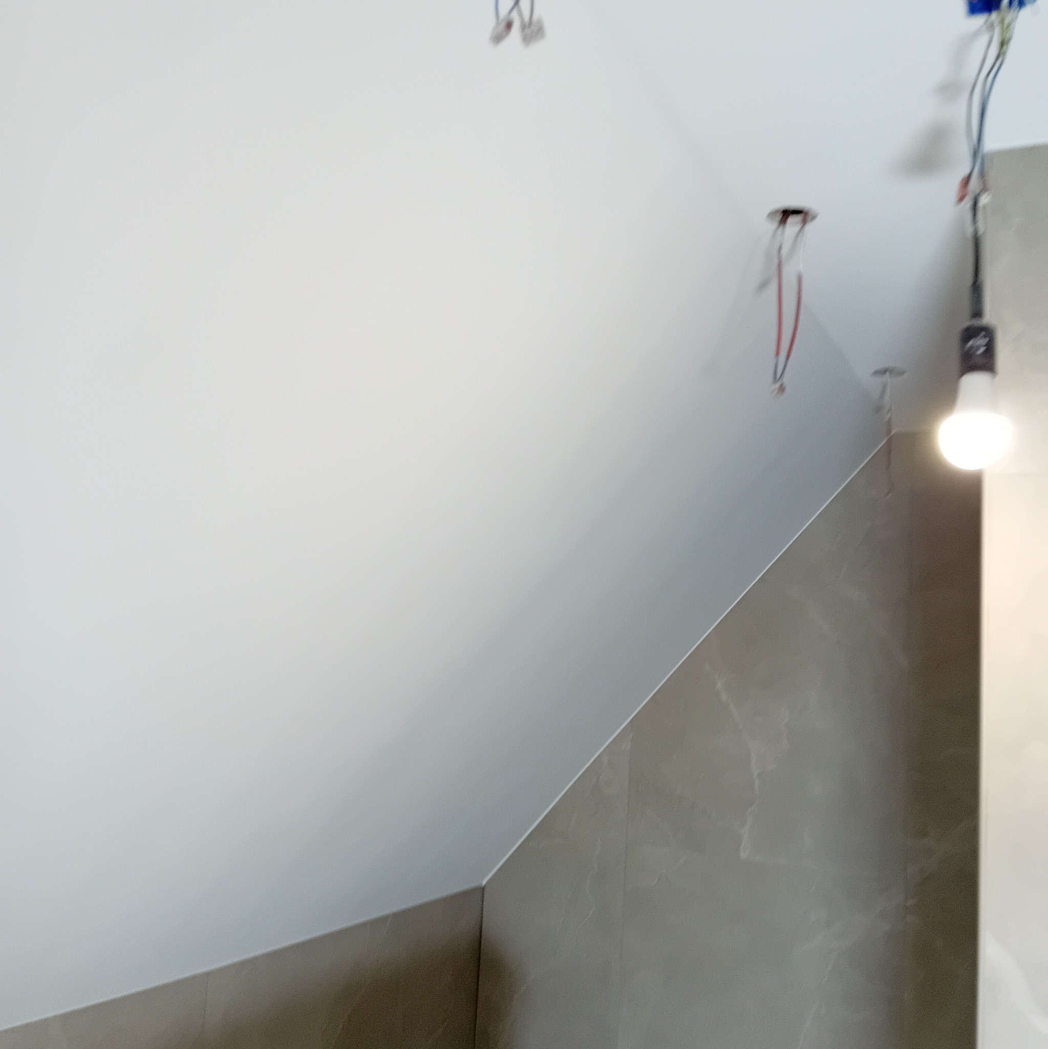 Skośny sufit połączony z szarą ścianą imitującą marmur, widoczne kable i otwory po oświetleniu, żarówka na kablu
