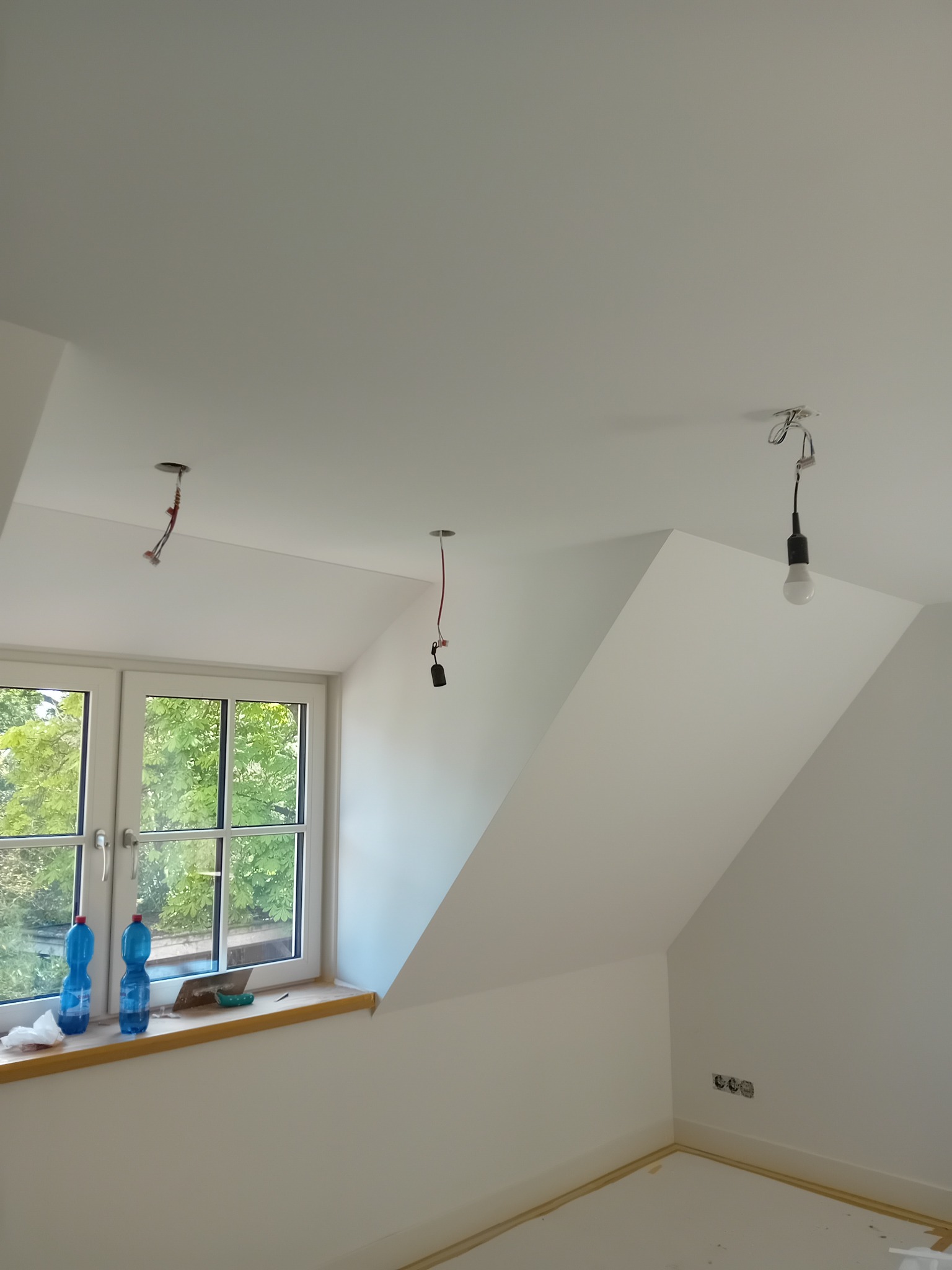 Poddasze w trakcie remontu: białe ściany, okno z widokiem na zieleń, odsłonięte kable oświetleniowe, zabezpieczona podłoga, butelki wody na parapecie.